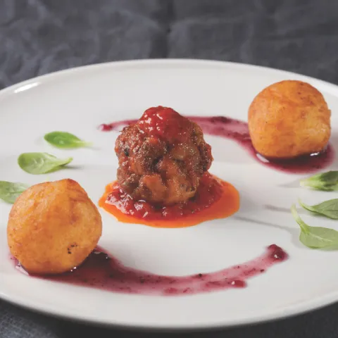Planeta de albóndiga de carne especiada sobre salsa marinera, acompañado de cristales de buñuelo de patata, ajo asado y queso crema, con estelas de salsa de arándanos y hojas de canónigos